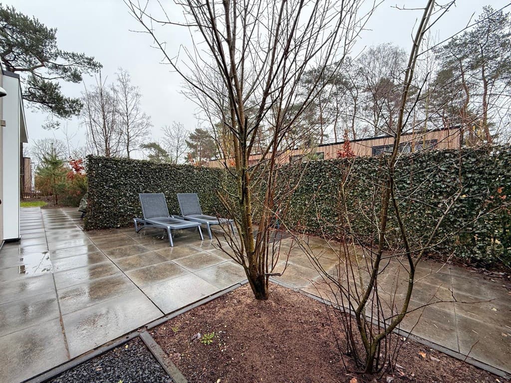 Terras met twee ligstoelen omringd door een groene haag en betegelde vloer, met bomen en planten in de omgeving