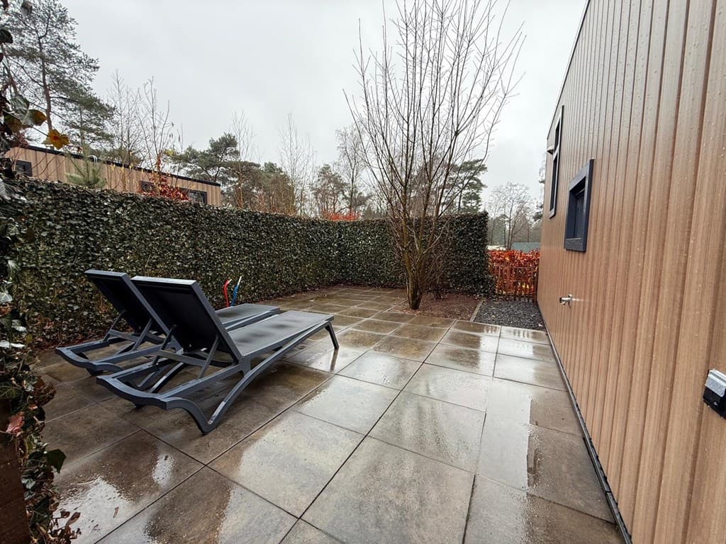 Terras met twee ligstoelen en een boom, omringd door hagen en met betegelde vloer in Ede.