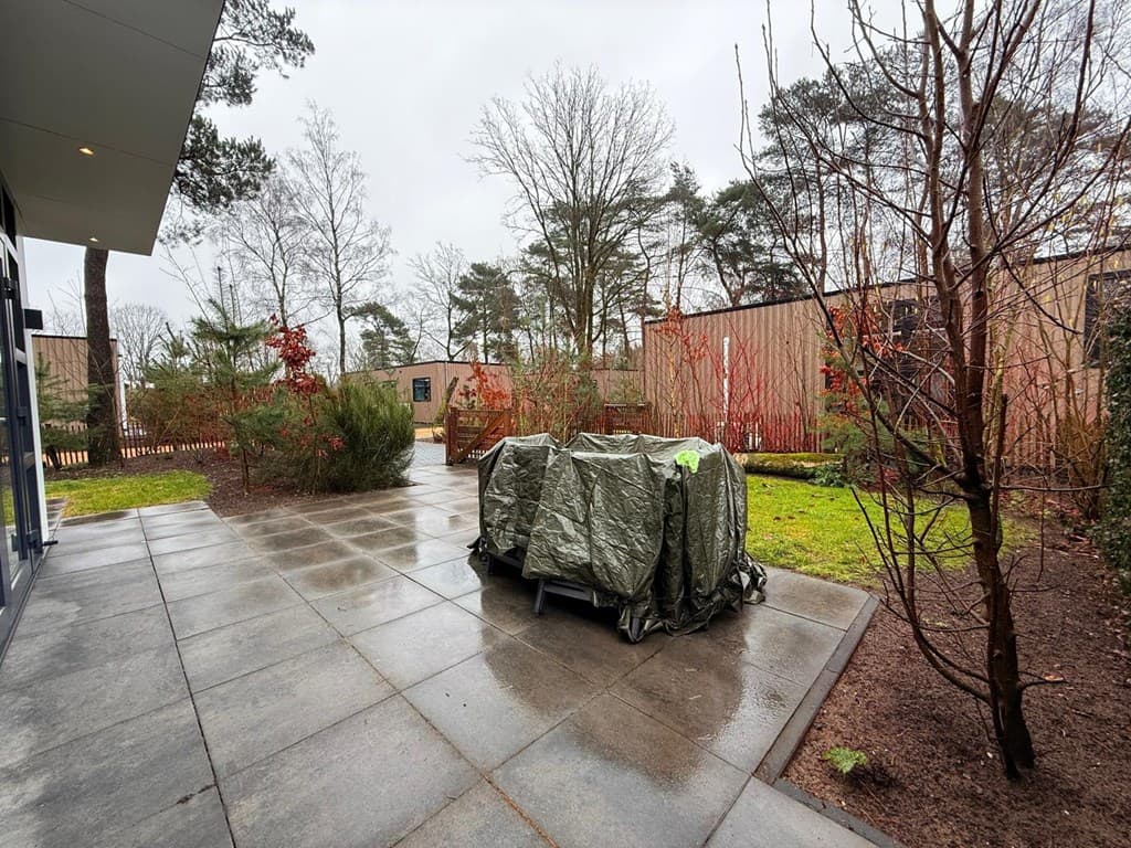 Tegels patio met een groen gebied en bedekte meubels, omringd door bomen en struiken, cloudy lucht