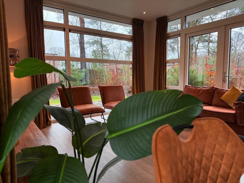 Lichte woonkamer met grote ramen, moderne meubels en uitzicht op de groene tuin met bomen en struiken.