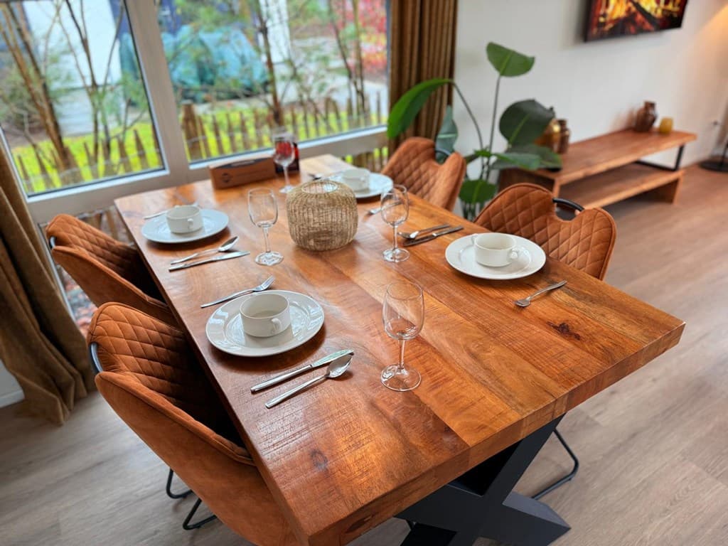 Eetkamer met een houten tafel, zes stoelen en gedekte tafel met borden, glazen en bestek, uitzicht op een groene omgeving.