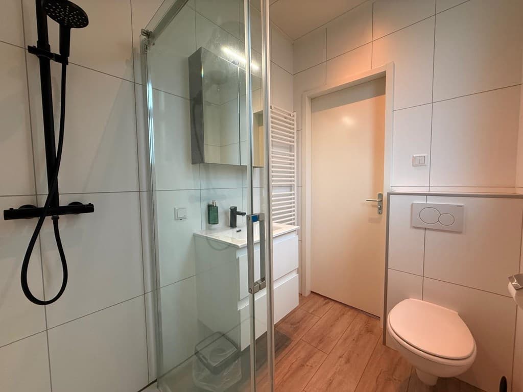 Moderne badkamer met inloopdouche, wastafel en toilet, witte tegelwand en houten vloer.