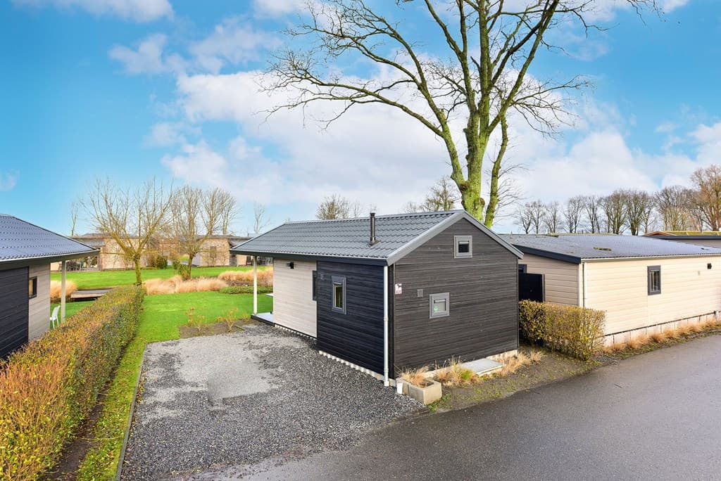 Zwart houten vakantiehuisje met grijze dakrand, omringd door groen gras en bomen in een vakantiepark