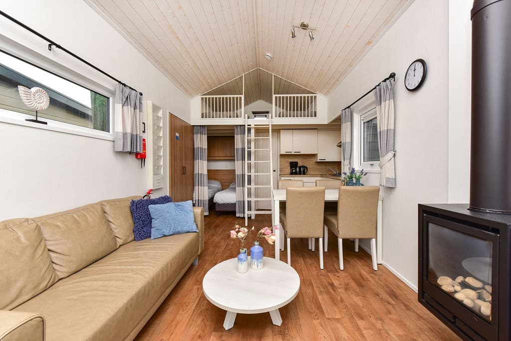 Ingerichte woonkamer met bank, eettafel en open keuken, bovenverdieping met twee bedden zichtbaar, houten vloeren en een kach