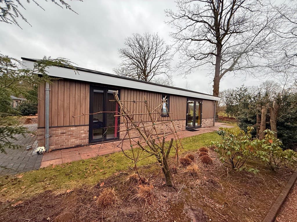 Moderne vakantiewoning met houten gevel, grote ramen en omringende tuin met gras en enkele bomen.