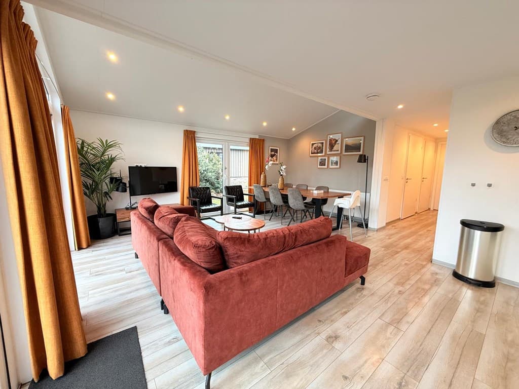 Moderne woonkamer met een grote rode bank, eettafel, televisie en grote ramen met gordijnen.