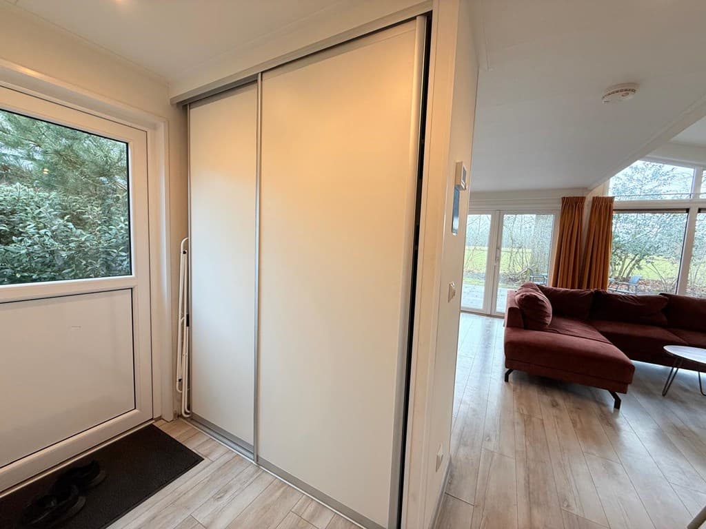 Ingang van de woning met een kast en een zicht op een woonkamer met een grote ramen en een zithoek met een rode bank.