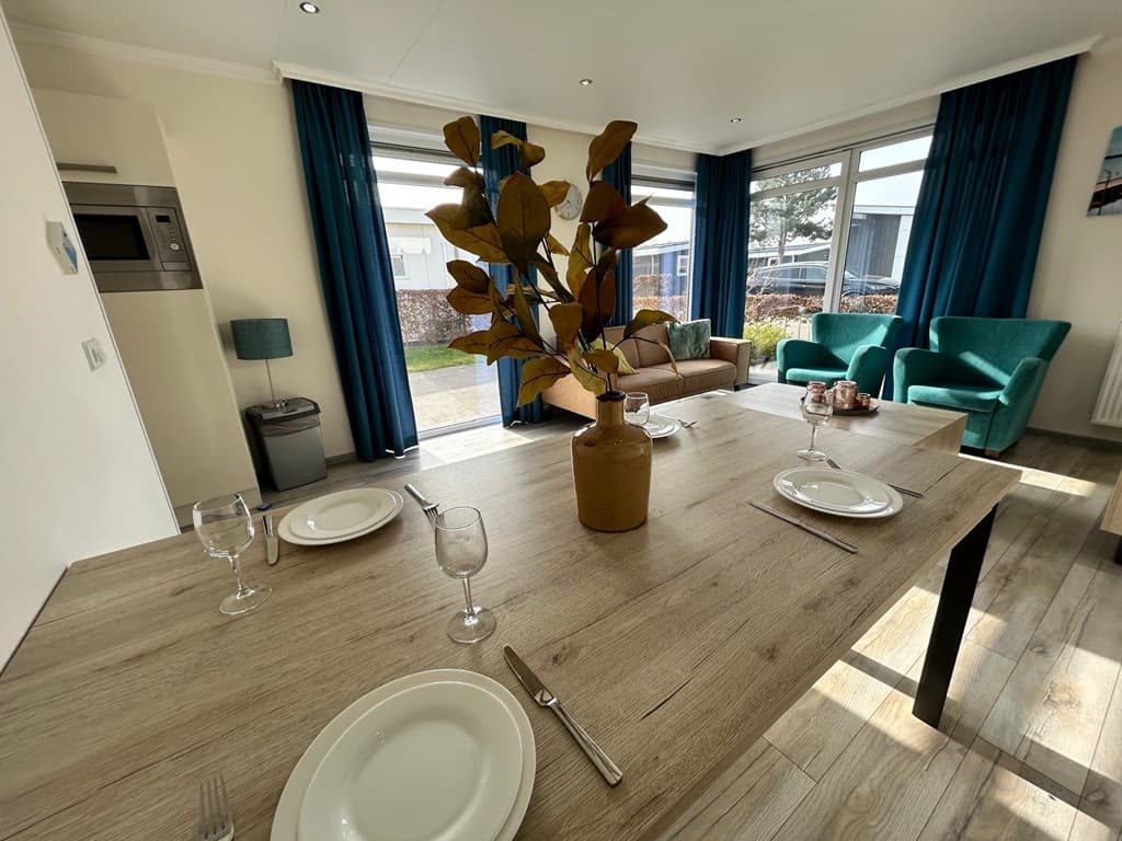 Eethoek met houten tafel, glazen op tafel, groene stoelen en een zithoek met bank en grote ramen met veel licht.