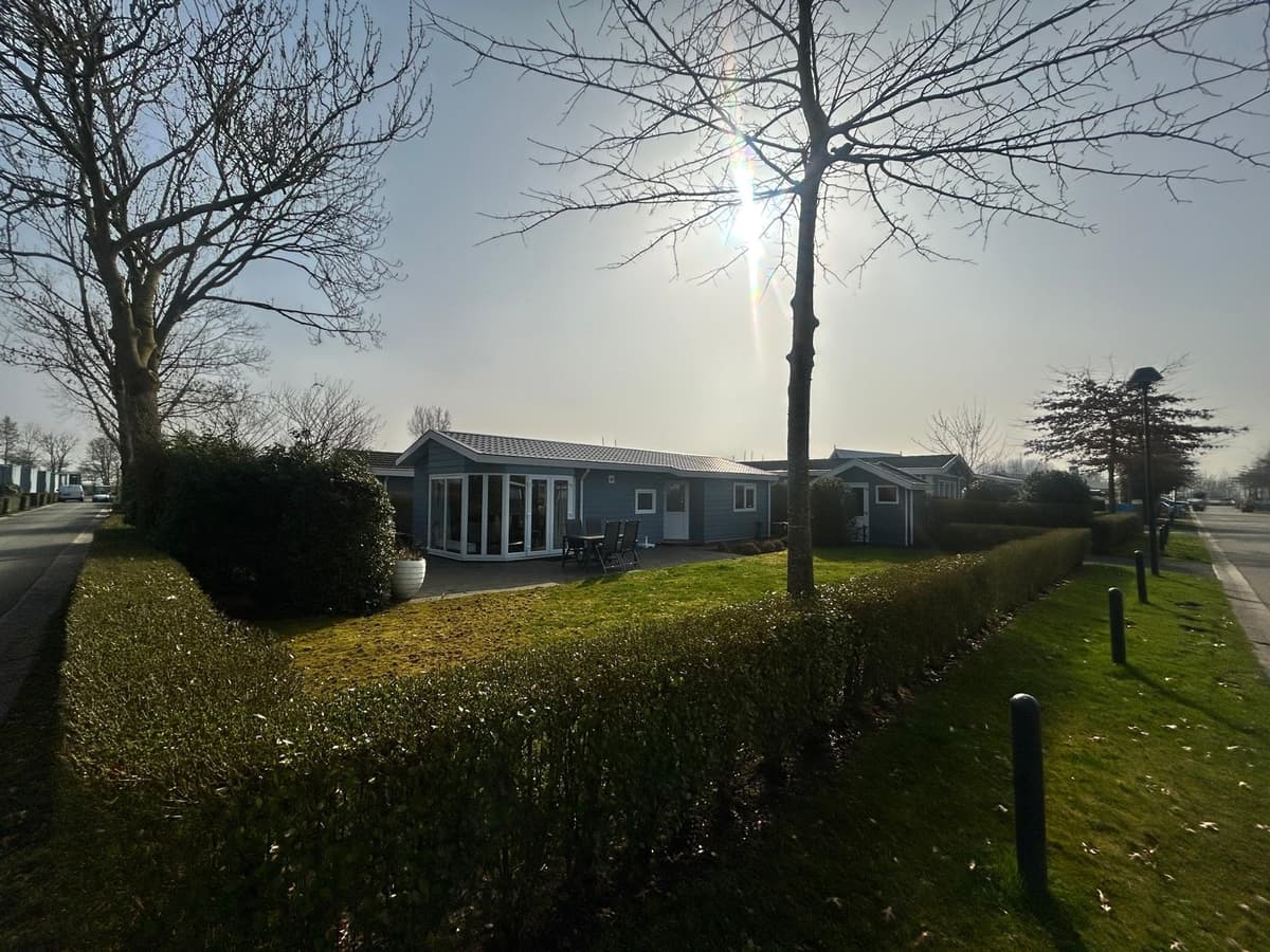 Buitenaanzicht van een vakantiewoning met groene tuin en terras, omringd door lage heggen en bomen, onder een zonnige lucht