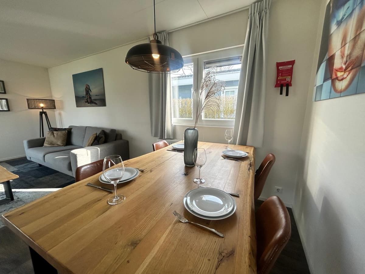 Eetruimte met houten tafel, stoelen, bord en bestek, naast een zithoek met een bank en wanddecoratie.