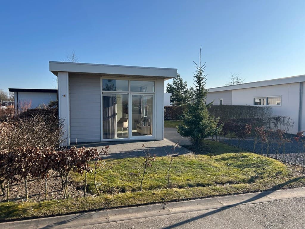 Moderne vakantiewoning met grote ramen en tuin, gelegen aan een verharde weg met groenblijvende planten.