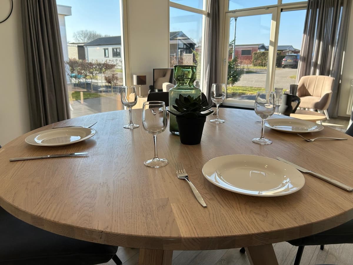 Eettafel met vier borden, glazen en een groene vaas in een lichte ruimte met grote ramen en uitzicht op buiten.