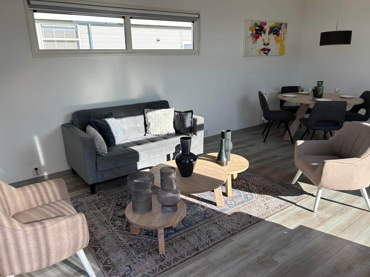 Zitgedeelte met een grijze bank, houten tafels, stijlvolle stoelen en kunst aan de muur in een lichte ruimte.