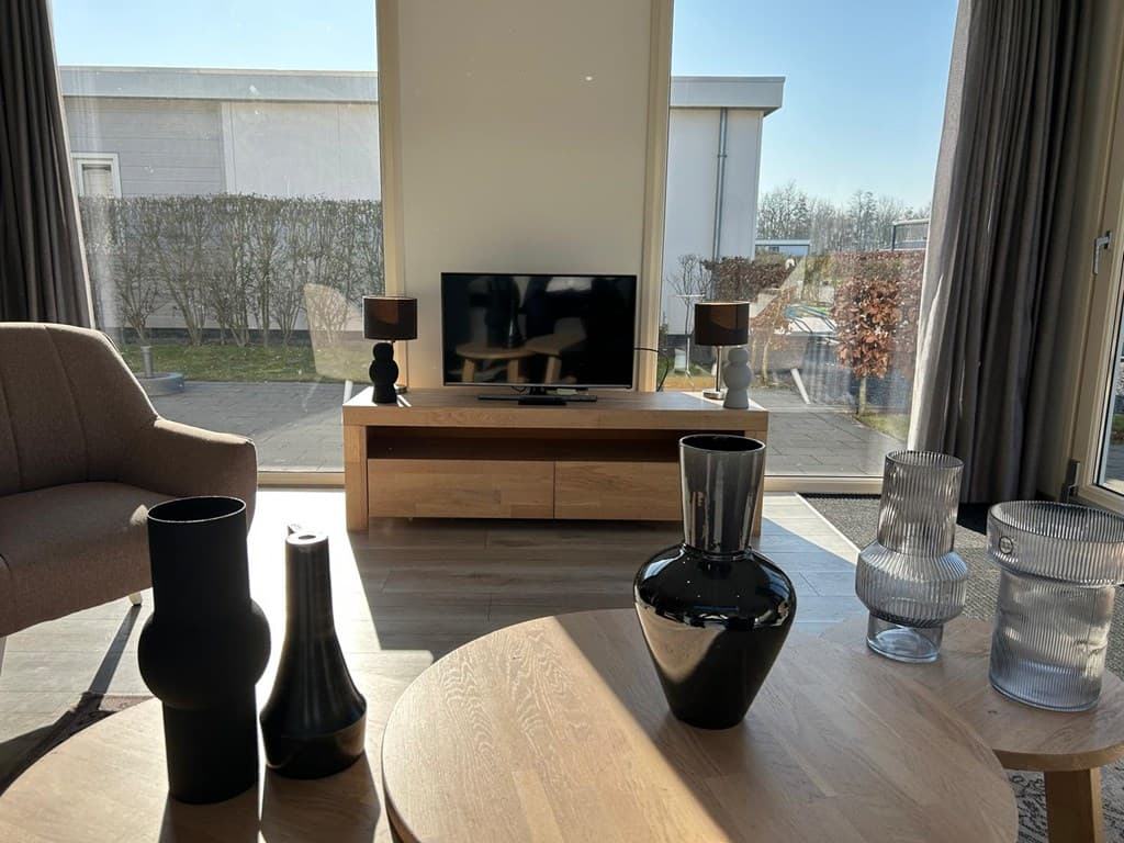 Lichte woonkamer met tv, moderne meubels en decoratieve vazen bij de ramen met uitzicht op de buitenruimte.