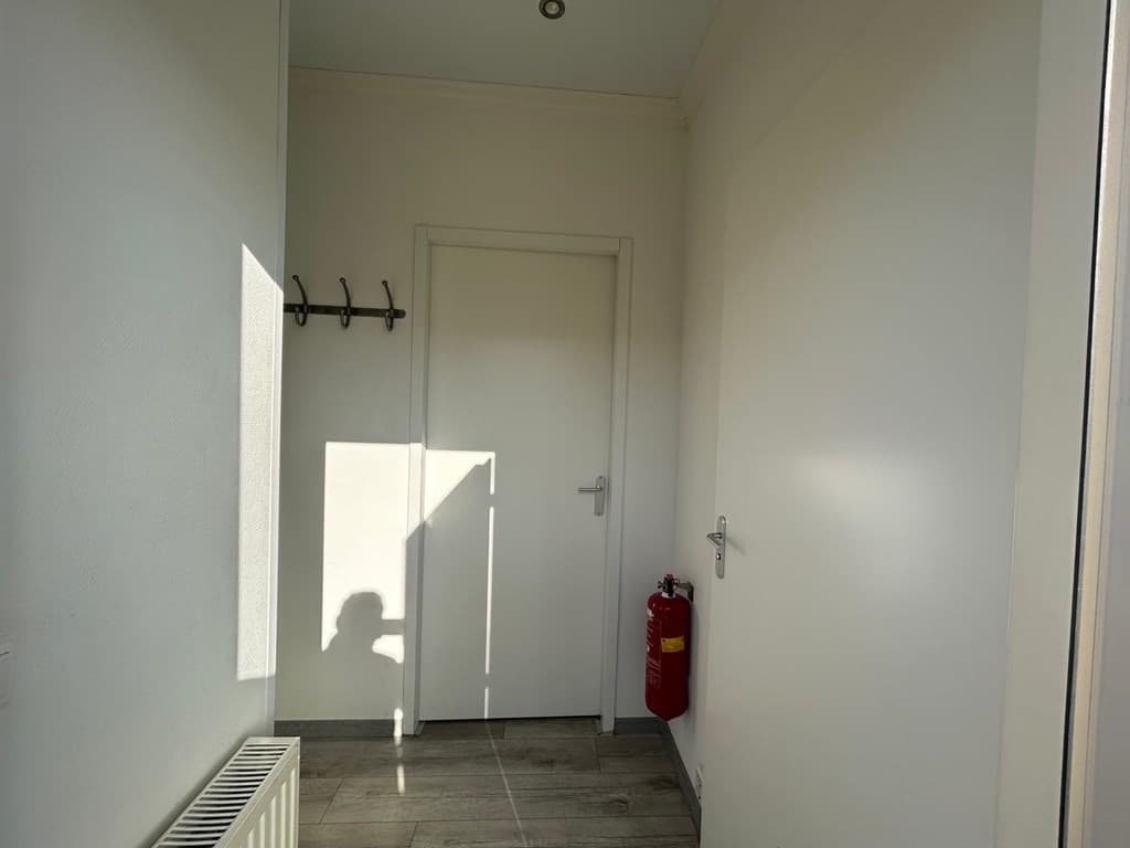Ingang van de woning met een deur, radiator, haakjes aan de muur en een brandblusser aan de zijkant.
