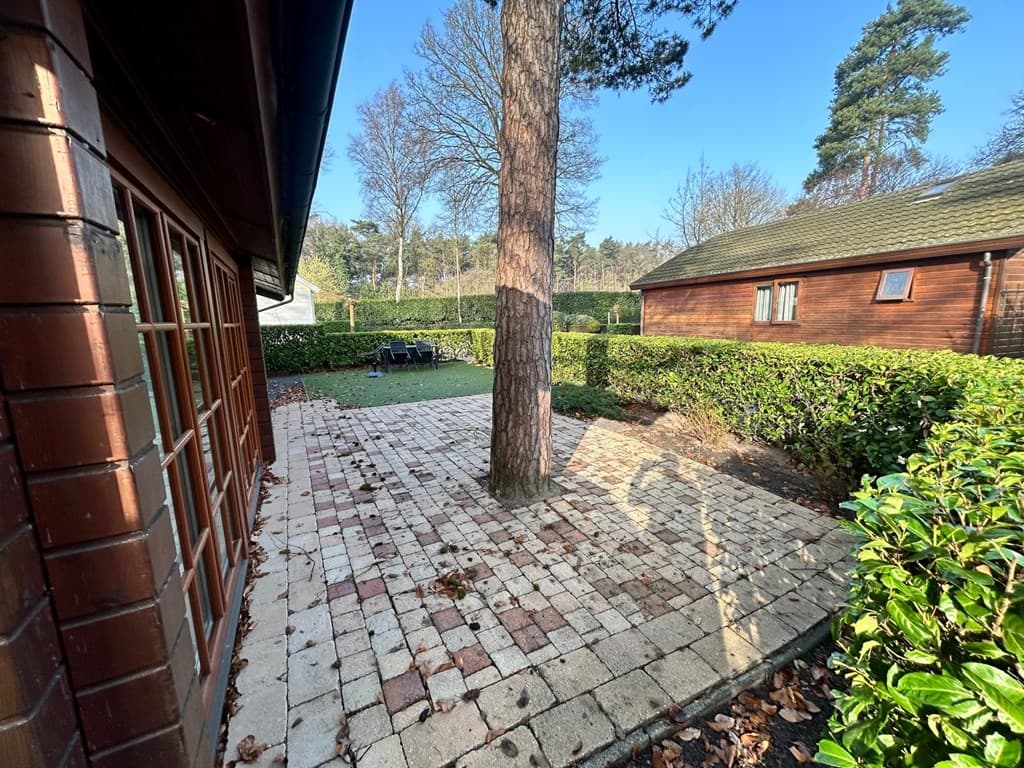 Buitenaanzicht van een vakantiewoning met een betegeld terras en een grote boom in de tuin, omringd door hekken.