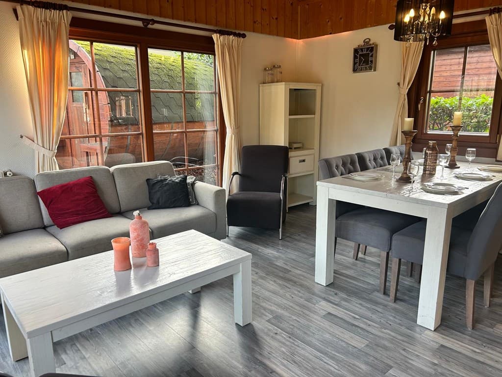 Moderne woonkamer met grijze bank, eettafel en uitzicht op een buitenruimte met een houten huisje en groene begroeiing.