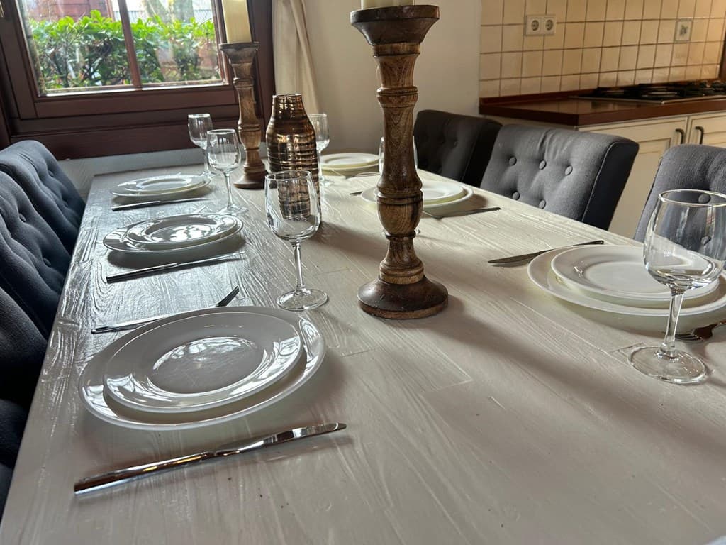 Houten eettafel gedekt met borden en glazen, met kaarsen en stoelen in een lichte, gezellige ruimte.