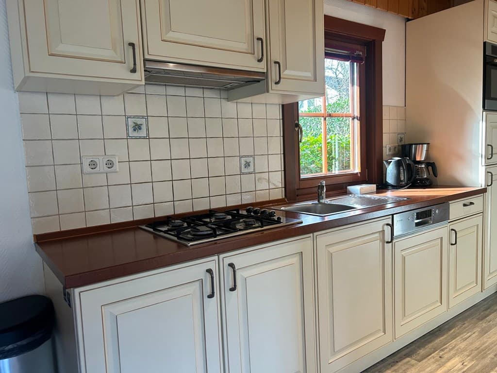 Keuken met lichte kastdeuren, gasfornuis, spoelbak en koffiezetapparaat bij een raam met uitzicht op de tuin.