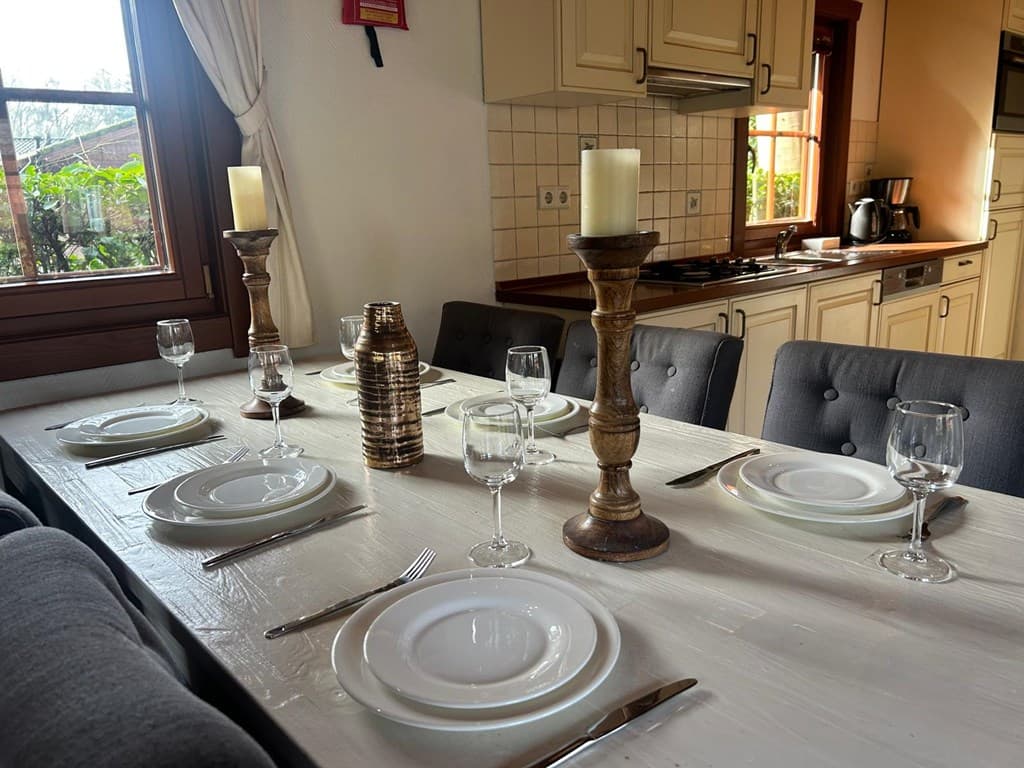 Eettafel met witte tafellaken, gedekte borden en glazen, naast een moderne keuken met houten kastjes en ramen.
