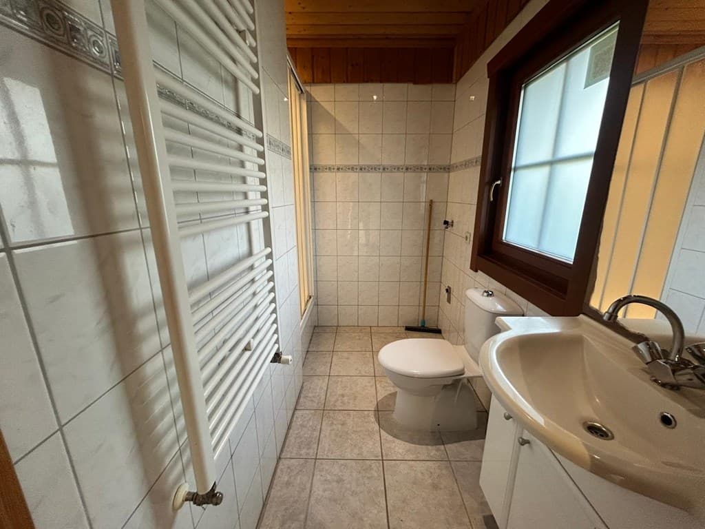 Badkamer met douche, toilet, wastafel en verwarming, betegelde wanden en vloeren in lichte kleuren.