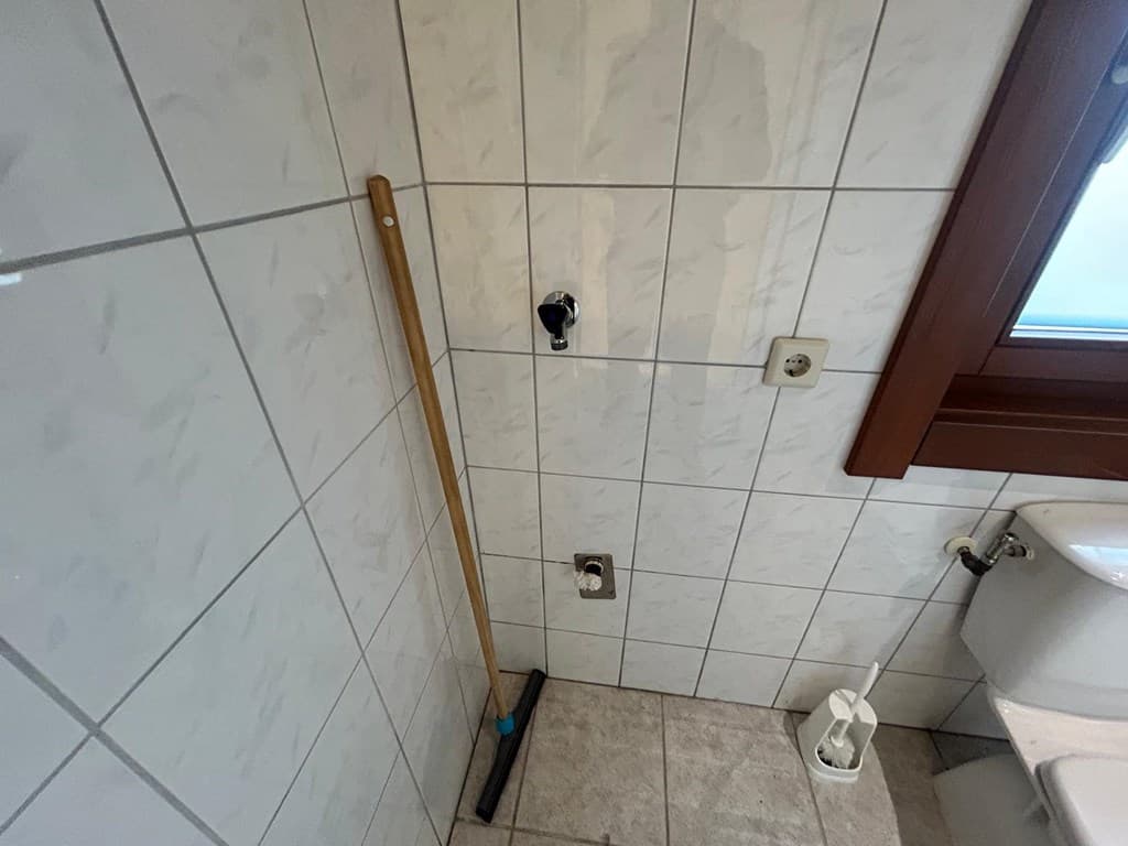 Een hoek van een badkamer met witte tegels, een raam en een schoonmaakstang naast de toiletpot.