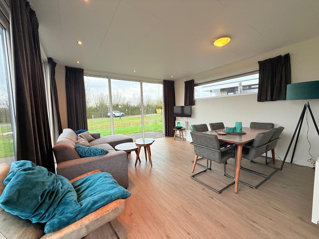Moderne woonkamer met grote ramen, bank, eettafel en stoelen, uitzicht op groen landschap en auto buiten.