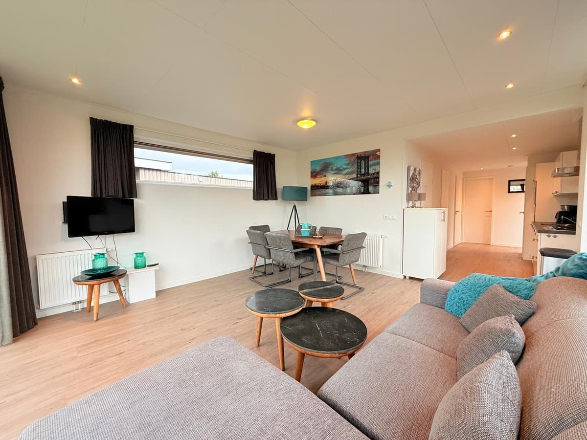 Lichte woonkamer met zithoek, eettafel en keuken, grote ramen en moderne inrichting.