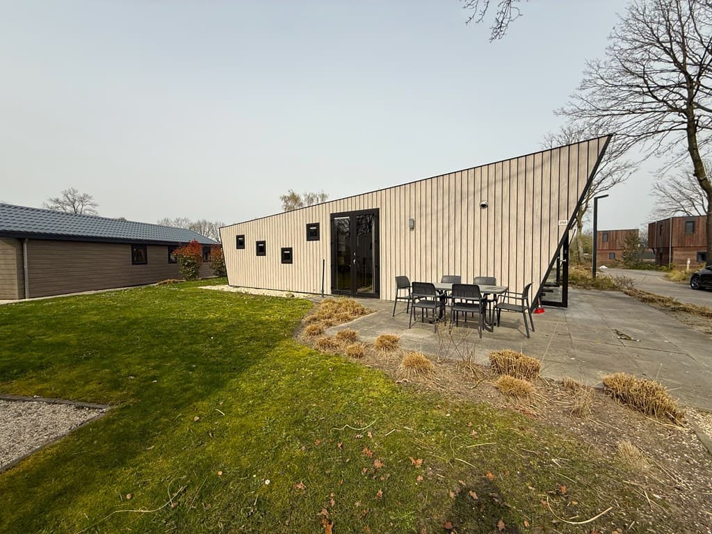 Moderne vakantiewoning met schuine gevel, terrasje met tafel en stoelen, groen gras en omliggende bomen.