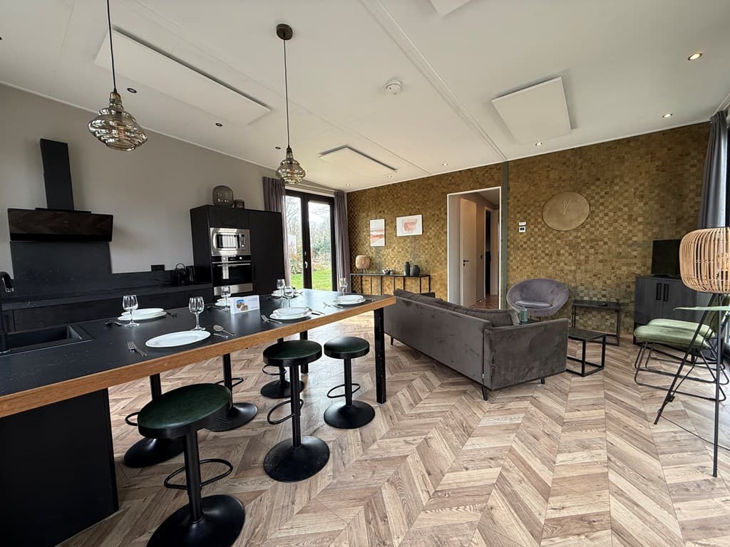 Moderne woonkamer met open keuken, houten vloer en eettafel met barkrukken, grote ramen en een groen accentmuur.