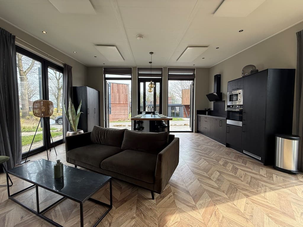 Moderne woonkamer met grote ramen, een bank, een eettafel en een keuken in donkere tinten, uitzicht op buitenruimte.