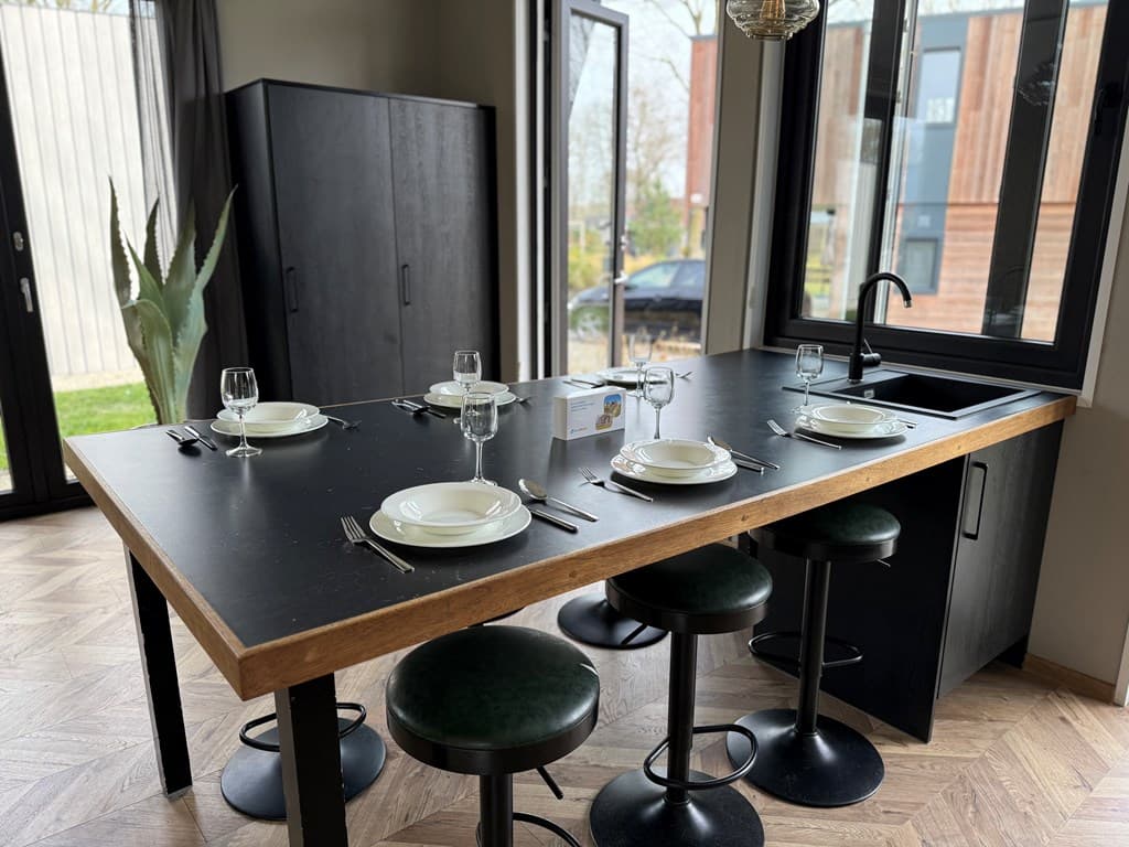Eetkamer met een houten tafel, vier borden en glazen, zwarte krukken en grote ramen met uitzicht naar buiten
