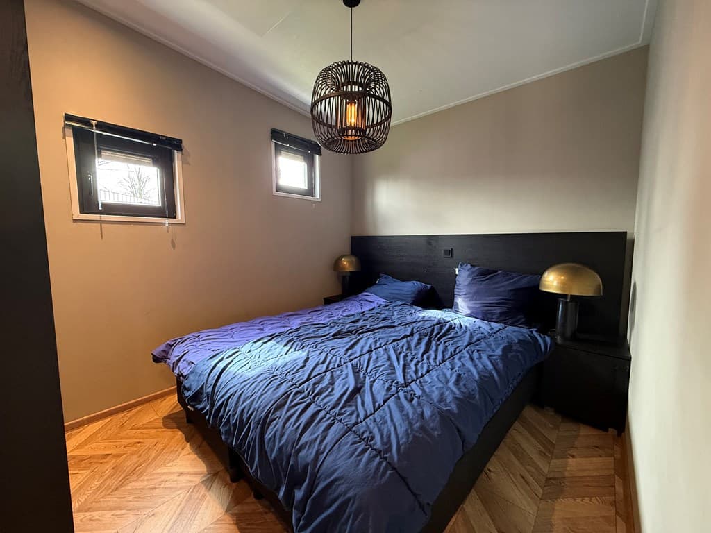 Sfeervolle slaapkamer met een tweepersoonsbed, nachtkastjes met lampen en kleine raampjes voor natuurlijk licht.