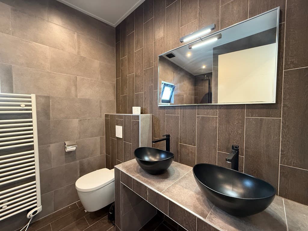 Moderne badkamer met grijze wandtegels, twee zwarte wastafels en een liggend raam voor natuurlijk licht.