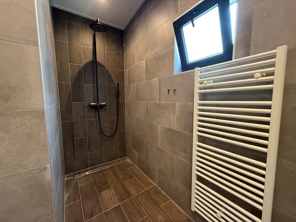 Moderne badkamer met inloopdouche, tegelwanden, en witte handdoekenradiator naast een klein raam