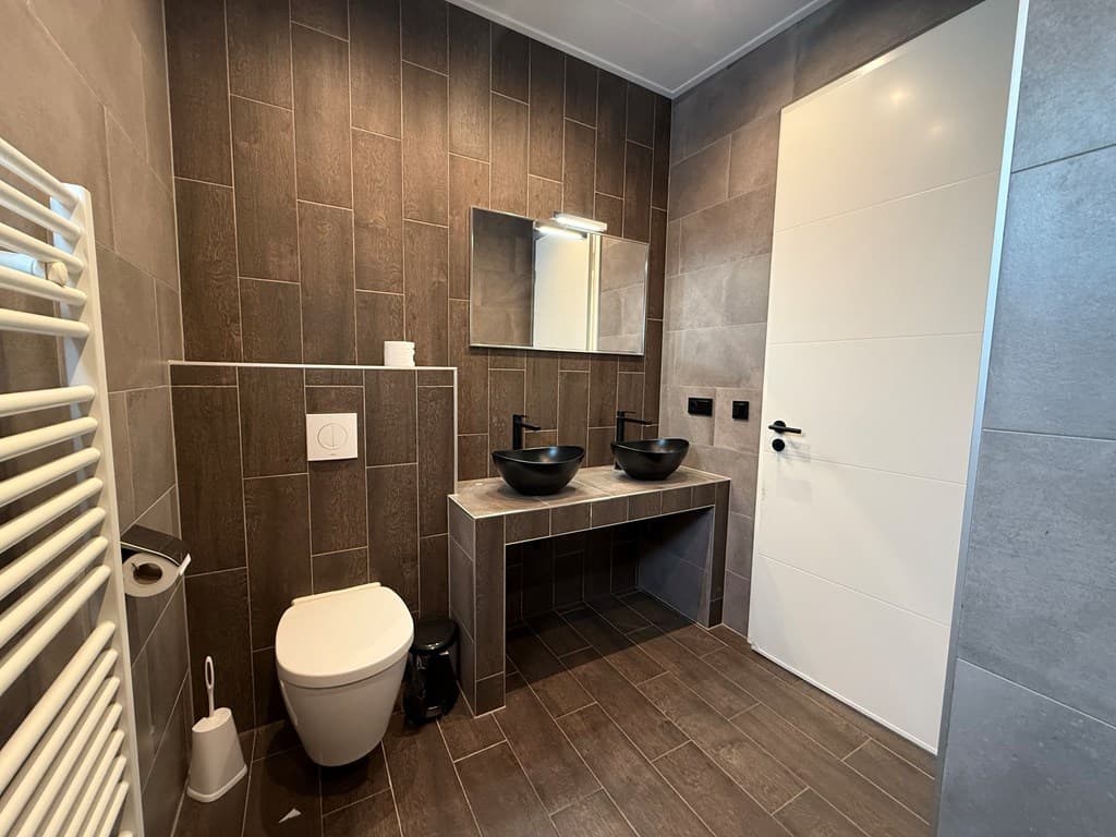 Moderne badkamer met twee zwarte wastafels, een toilet en een handdoekradiator op donkerbruine tegels