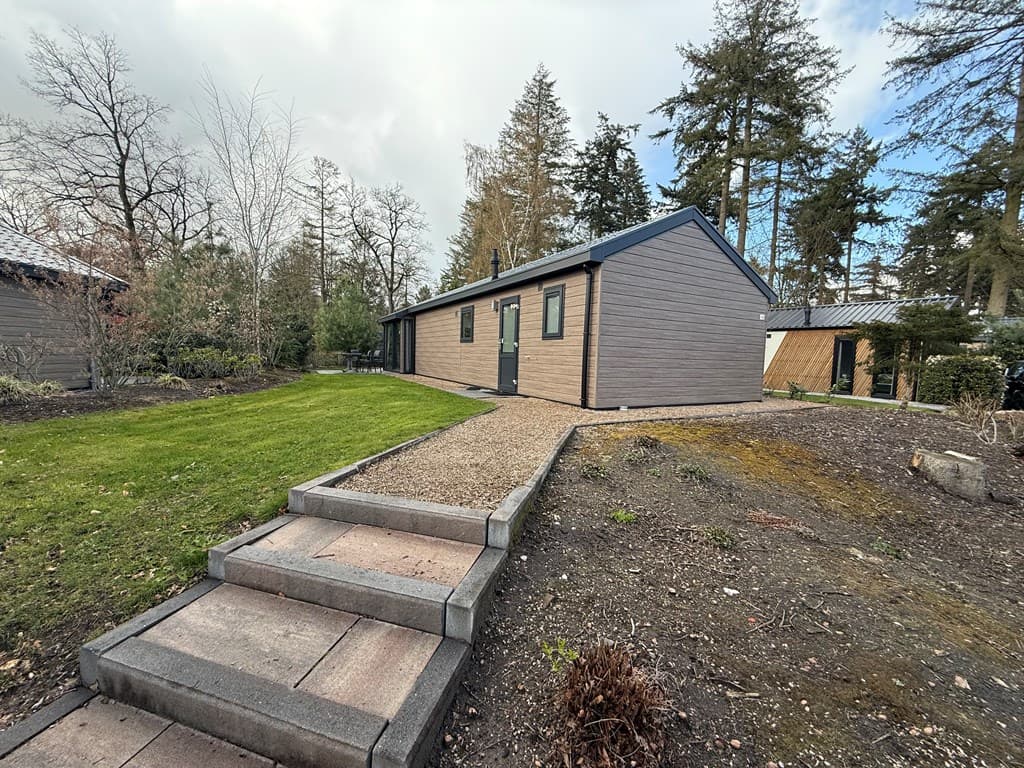Moderne vakantiewoning met een grijze en beige buitenkant, omgeven door gras en bomen, en een stijle trap naar de entree.