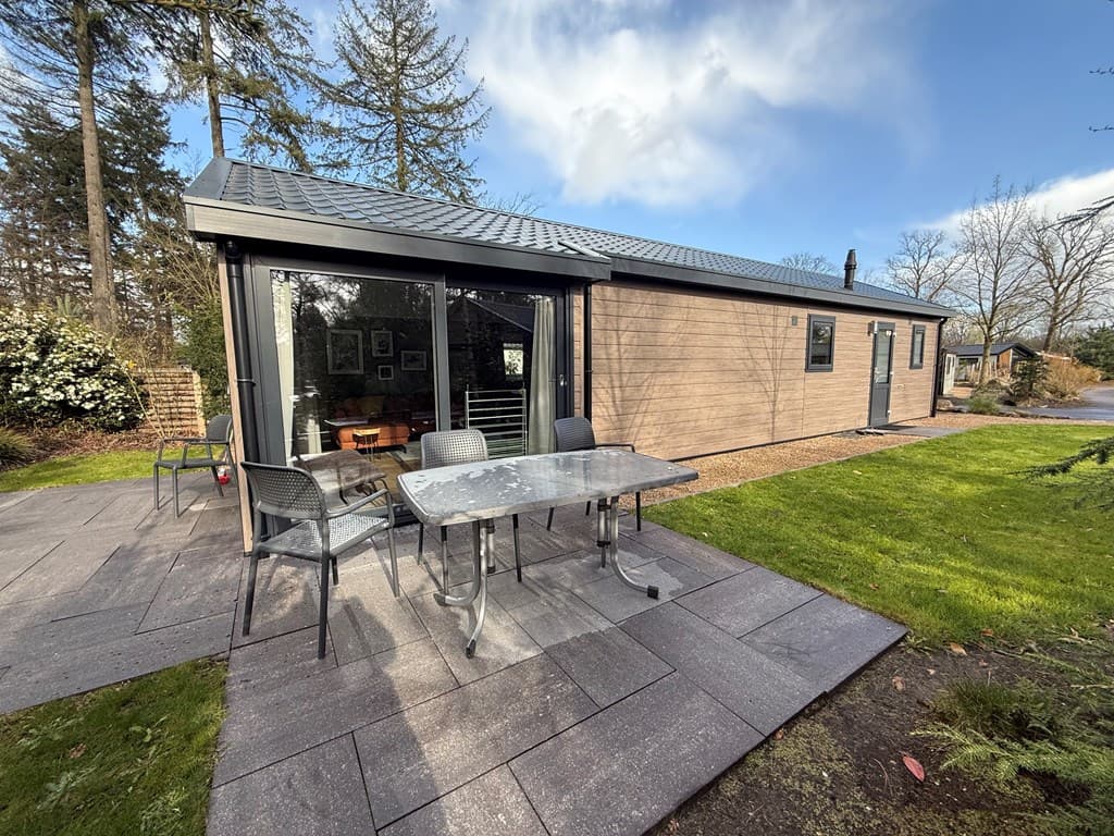 Moderne vakantiewoning met een terras, tuinmeubels en een betegeld pad, omgeven door bomen en groen.