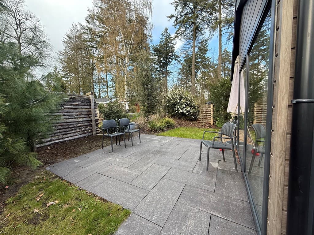 Terras met vloer van tegels, twee stoelen en tafeltje, omringd door groene bomen en struiken