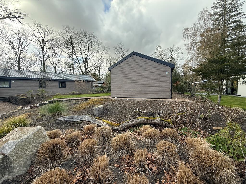 Zijaanzicht van een moderne vakantiewoning omringd door bomen en een aangelegde tuin met gras en heesters