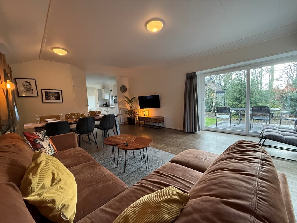Interieur van een woonkamer met een bruine bank, een salontafel, en een open verbinding naar de eethoek en keuken met uitzich