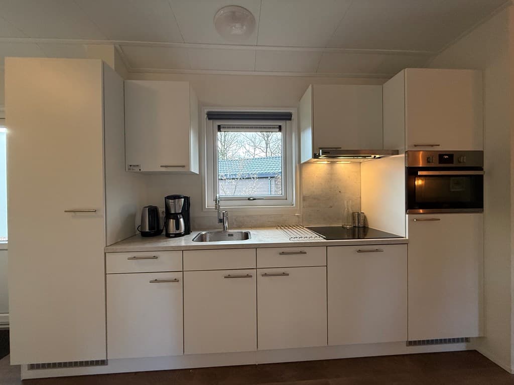 Moderne keuken met witte kasten, fornuis, oven, en een klein aanrecht met spoelbak en koffiezetapparaat.