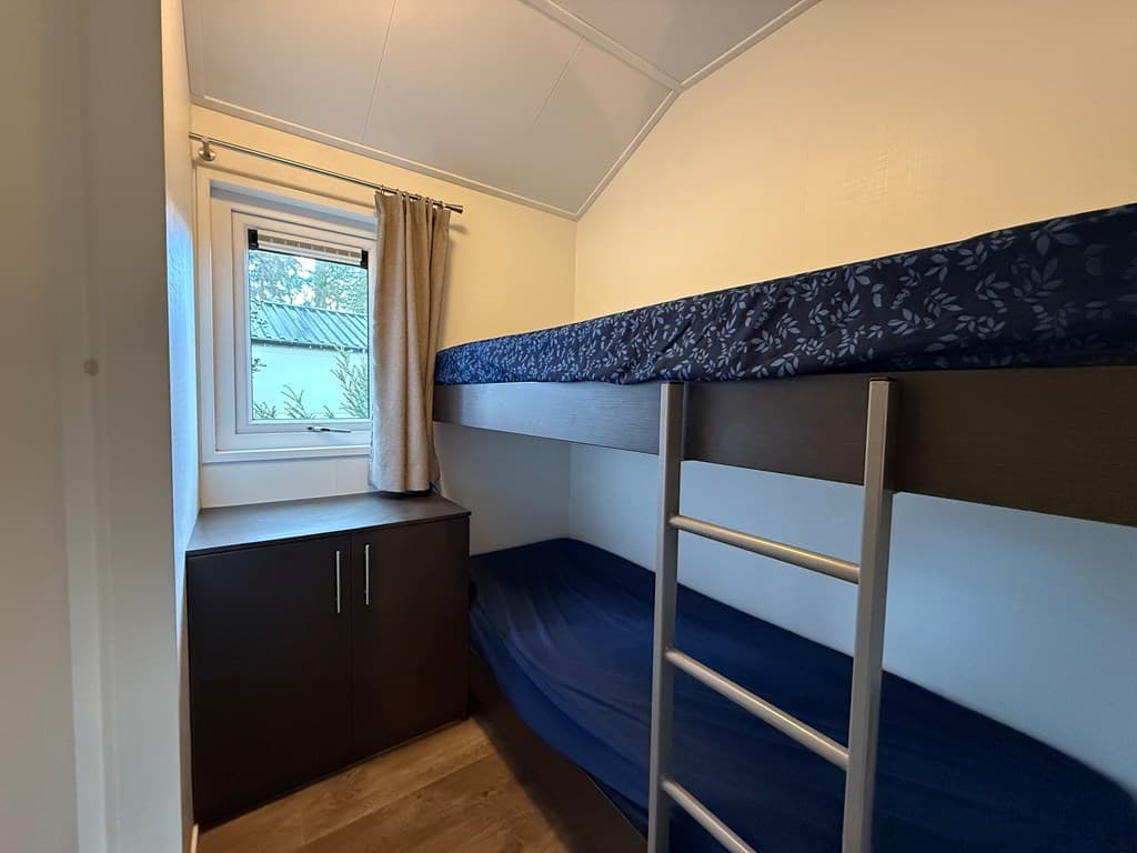 Slaapkamer met stapelbed en kast, voorzien van een raam met lichte gordijnen.
