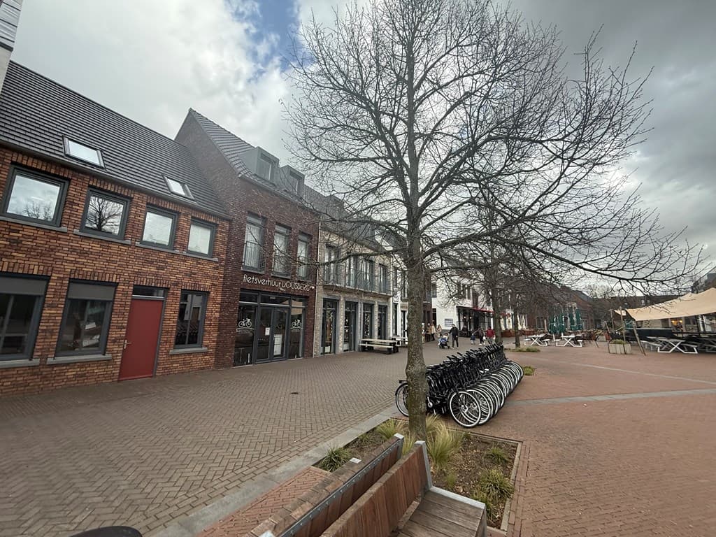 Straatbeeld met gebouwen en een boom, fietsen geparkeerd langs de stoep en terrassen in de verte.