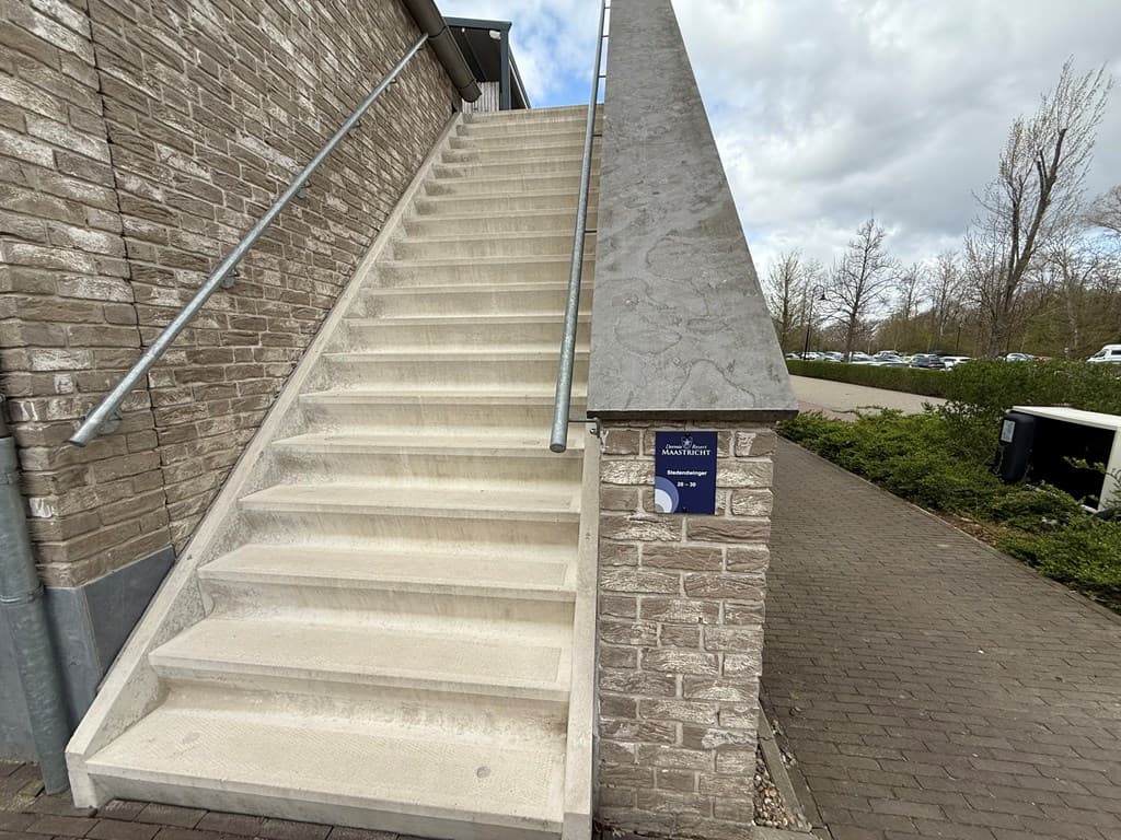 Trap met schuine wand en bordje bij Dormio Resort Maastricht, omgeven door groen en parkeerplaatsen in de achtergrond