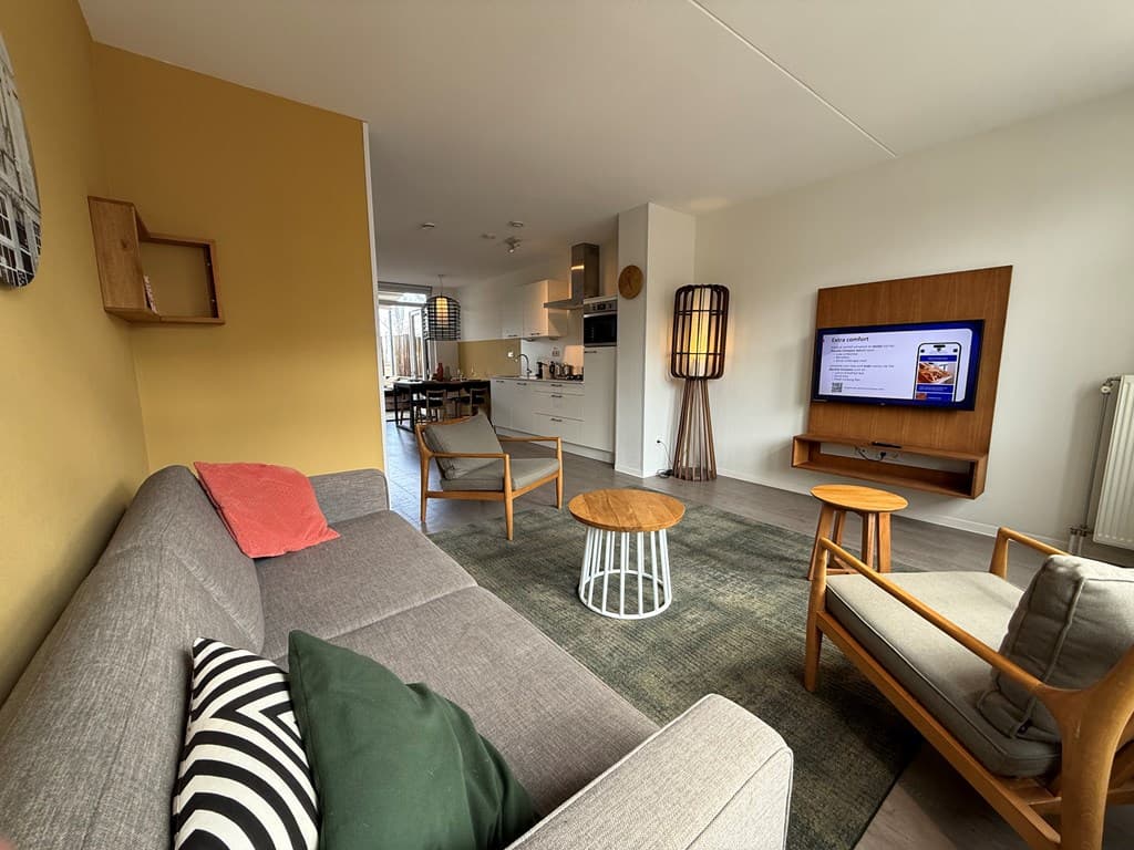 Modern uitgeruste woonkamer met zithoek, televisie en toegang tot de open keuken in lichte kleuren.