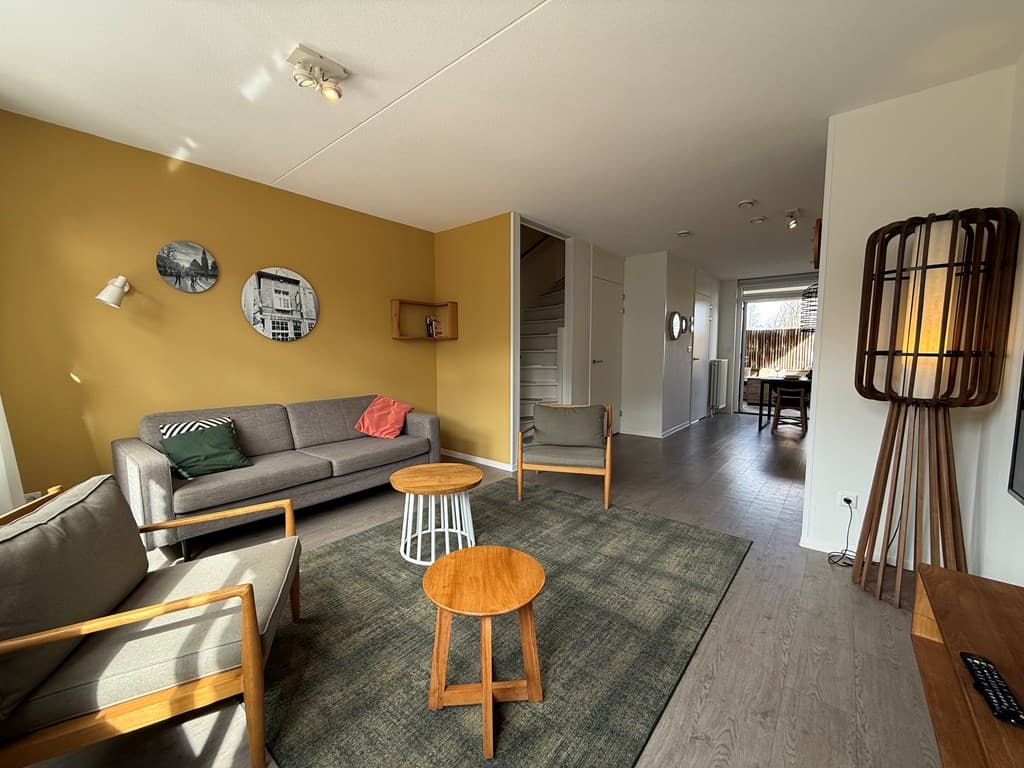 Woonkamer met een gele muur, moderne meubels, een vloerkleed en toegang tot de eetruimte en trap naar boven