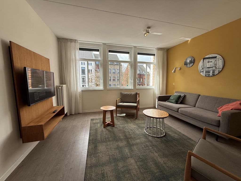Woonkamer met grijze bank, houten meubels en grote ramen met uitzicht op een straat in de stad.