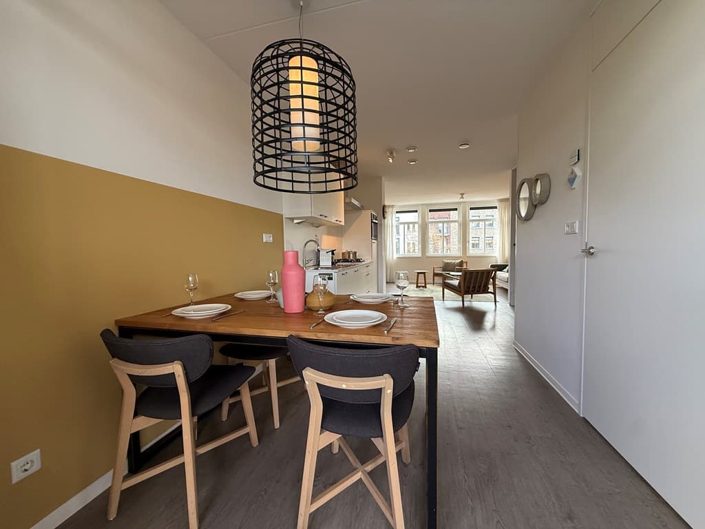 Eetkamer met houten tafel, zwarte stoelen, lamp en toegang naar een lichte woonkamer met grote ramen en moderne inrichting