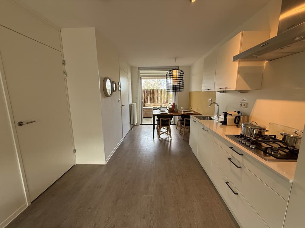 Lichte open keuken met moderne apparatuur en een eetruimte met een grote raam en uitzicht op de buitenruimte.